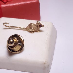 May include: Gold-toned jewelry set featuring a mouse-shaped pin with a red gemstone eye and a round, layered pin. The set is displayed on a white surface with a red gift box in the background. A unique accessory.