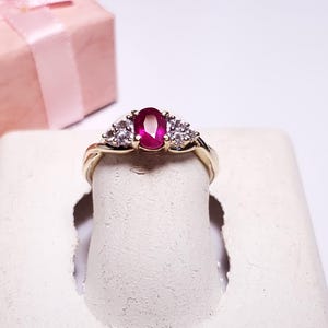 May include: A gold ring with a large oval ruby gemstone and smaller diamond accents. The ring is displayed on a white ring holder.