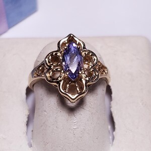 May include: A gold-toned ring with a marquise-cut, light purple gemstone at the center. The ring features an openwork design with floral-inspired details. The ring is displayed on a white surface, with a blue box in the background.