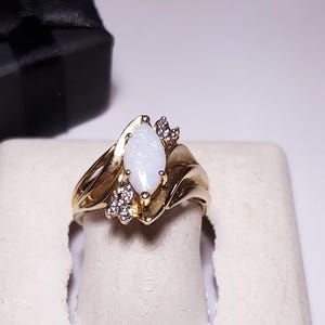 May include: A gold ring with a marquise-cut opal center stone, flanked by small diamond accents. The ring is set on a white display stand, with a black gift box in the background. The ring's design features a swirling, elegant shape.