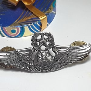 May include: A silver-toned military pilot wings pin with a star and wreath design. The wings are spread and the pin has a safety clasp.