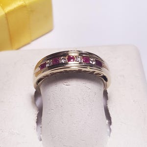 May include: A gold-colored ring with a row of alternating red and clear stones. The ring is displayed on a white surface, with a yellow gift box in the background. The ring is a band style.