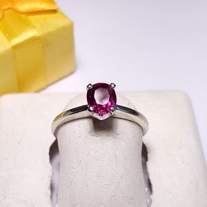 May include: A silver ring with a vibrant pink oval gemstone. The ring is displayed on a white stand, with a yellow gift box with a bow in the background. The ring is a piece of jewelry.