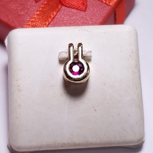 May include: A gold pendant featuring a single round, faceted, deep pink gemstone set in a circular bezel. The pendant has a vertical bail at the top and is displayed on a square white card, with a red gift box visible in the background.