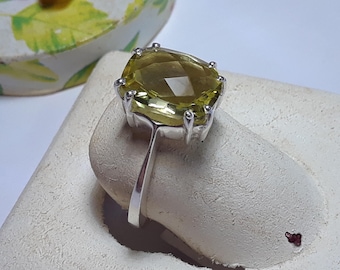 May include: A silver ring with a large, faceted, yellow-green gemstone set in a square-shaped bezel. The ring is on a white display stand.