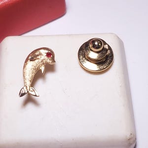May include: A gold-toned dolphin pin with a red gem eye and a matching gold-toned tie tack. The dolphin pin is detailed with fins and a tail. The items are displayed on a white surface, with a red gift box in the background.