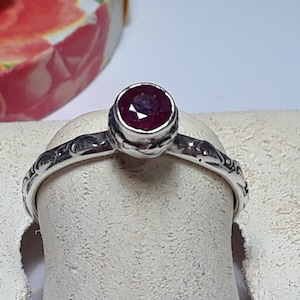 May include: A silver ring with a red gemstone set in a bezel setting. The band is decorated with a delicate floral design.