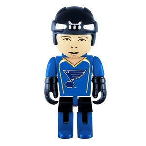 May include: A blue and yellow hockey player figurine wearing a black helmet with a white stripe. The figurine is wearing a blue jersey with a yellow bird logo and the word "Blues" on the front. The figurine is standing on a white background.