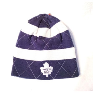 May include: A blue and white striped knit beanie with a quilted diamond pattern. The beanie has a white embroidered Toronto Maple Leafs logo.