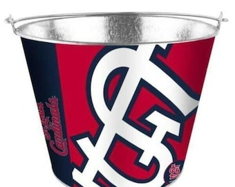 Puede incluir: Un cubo de hielo de metal rojo, blanco y azul con el logotipo de los St. Louis Cardinals. El cubo tiene un asa y es perfecto para mantener las bebidas frías en una fiesta o un juego.