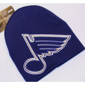 May include: A navy blue knit beanie with a white and yellow embroidered St. Louis Blues logo. The logo features a stylized musical note with wings.