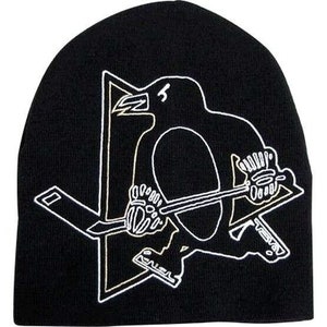 May include: Black knit beanie with a white outline of a penguin holding a hockey stick. The penguin is inside a star shape.