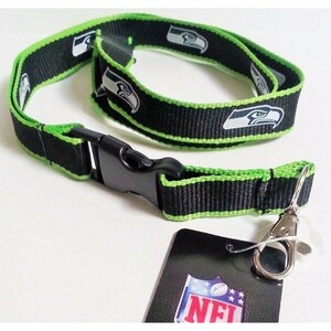 May include: Black and green lanyard with a green and white Seattle Seahawks logo. The lanyard has a black plastic clip and a silver metal hook. The lanyard is packaged with a black and white NFL logo tag.