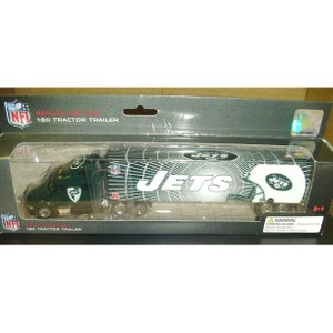 May include: A green and white New York Jets tractor trailer replica. The trailer features the team's logo and the word "JETS" in large letters. The packaging includes the NFL logo and the text "1:80 Tractor Trailer".