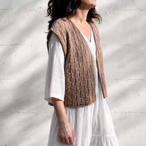 Può includere: Un gilet lavorato a maglia beige con motivo a trama aperta. Il gilet senza maniche è indossato sopra un vestito bianco. Lo sfondo è un muro bianco con ombre.