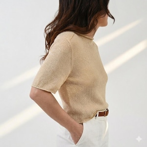 Puede incluir: Top de punto beige de manga corta y cuello alto. El top se combina con pantalones blancos y un cinturón de cuero marrón con hebilla dorada. La modelo está de pie frente a un fondo blanco.