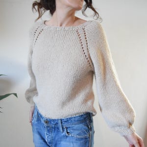Peut inclure: Un pull en tricot beige avec un col rond et des manches longues. Le pull présente un délicat motif de dentelle sur les épaules et les poignets.