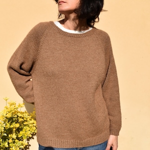 May include: A brown knitted jumper with a crew neck and raglan sleeves. The jumper is a solid colour and appears to be made of a soft, warm material. The jumper is worn over a white t-shirt.
