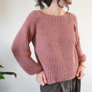May include: A pink knitted sweater with a ribbed texture and a slightly oversized fit. The sweater has a boat neck and long sleeves.