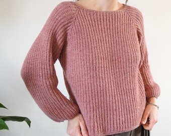 Può includere: Un maglione di lana rosa con una trama a coste e una vestibilità leggermente oversize. Il maglione ha un collo a barca e maniche lunghe.