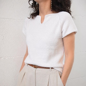 May include: A white knit short-sleeve top with a notched neckline. The top is paired with tan pants and a brown belt with a gold-colored buckle. The outfit is simple and casual.