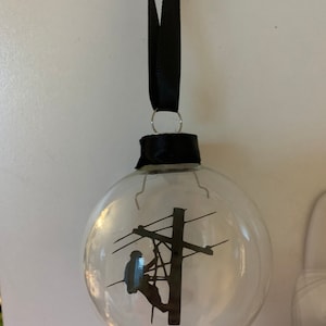 May include: A clear glass ornament with a black silhouette of a person working on a power line.