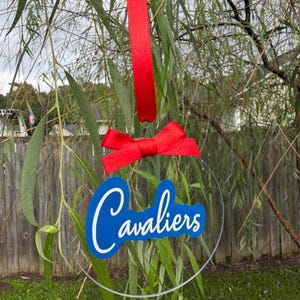 May include: A circular clear acrylic ornament featuring the word Cavaliers in white script lettering on a blue background. The ornament is suspended by a red ribbon and includes a decorative red ribbon bow at the top of the clear circular disc.