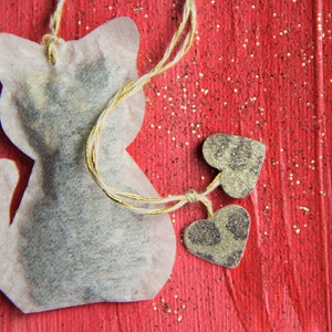 May include: A white tea bag shaped like a cat with two small silver hearts attached to it. The tea bag is on a red background with gold glitter.