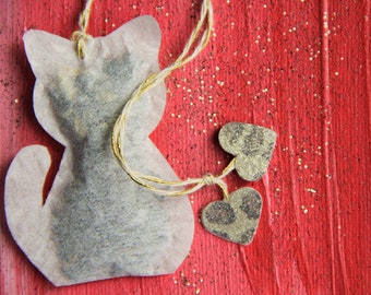 May include: A white tea bag shaped like a cat with two small silver hearts attached to it. The tea bag is on a red background with gold glitter.
