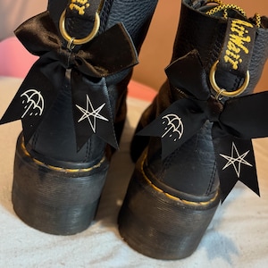 May include: Black leather boots with chunky soles, adorned with black bows displaying white graphic prints. The boots have yellow stitching and a yellow and black pull tab.