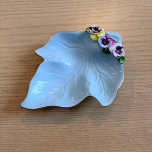 May include: A light blue, leaf-shaped ceramic dish with gold trim. The dish is decorated with small, colorful pansy flowers in shades of purple, yellow, and white. The dish is a decorative item.