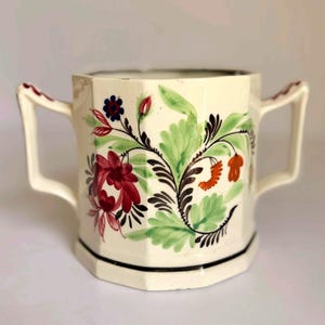 May include: An off-white ceramic sugar bowl with an octagonal shape and two side handles. The exterior features a hand-painted floral pattern in shades of red, green, and dark blue, with a thin black line accenting the base of the vessel.