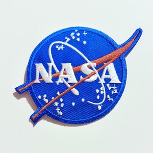 May include: A circular embroidered patch featuring the blue NASA logo with white lettering. The design includes a red orbital swoosh crossing the text, surrounded by white star-like dots on a blue background with a blue stitched border.