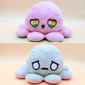 May include: A reversible plush octopus toy featuring two distinct sides. One side is pink with embroidered heart-shaped eyes containing rainbow stripes and a smiling mouth. The other side is light gray with embroidered sad, downturned eyes and a wavy mouth.