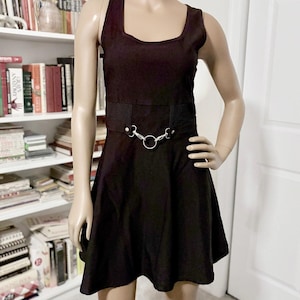 May include: A sleeveless black dress featuring a square neckline and a flared skirt. The waist is accented with a horizontal mesh panel and a silver-toned metal ring hardware detail connected by two small silver clips at the center front.