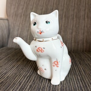 May include: A white ceramic teapot shaped like a sitting cat with one paw raised. The teapot features a gold collar, painted green eyes, and small orange and black floral patterns across its body. The cat's head serves as the lid for the teapot.
