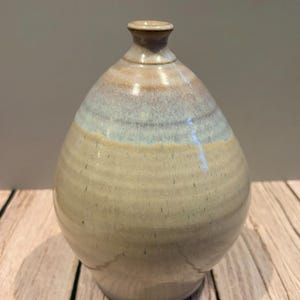 May include: A ceramic vase with a rounded, bulbous body and a narrow neck. The surface features a smooth, glossy glaze with a color gradient transitioning from a light, sandy beige at the base to a soft, muted blue hue near the top rim.