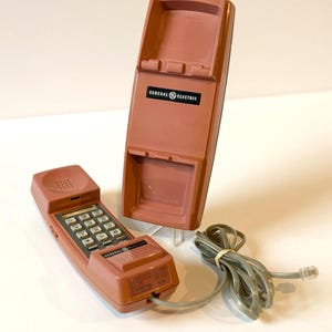 May include: A dusty rose-colored General Electric wall-mount telephone with a matching handset. The unit features a rectangular base with a black logo plate, a numeric keypad, and a coiled gray cord with a standard modular telephone connector plug.