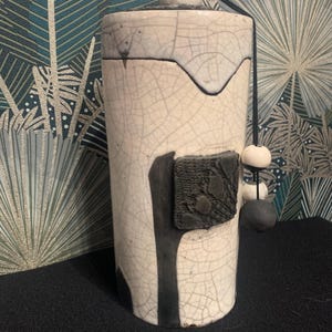 Peut inclure: Un récipient cylindrique en céramique avec un couvercle, présentant un glaçage craquelé blanc et des accents noirs. Une bande verticale noire et un carré texturé ornent le côté. Un cordon noir avec des perles blanches et noires pend du couvercle.