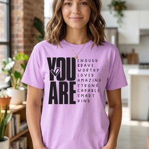 May include: A light purple t-shirt with the words "YOU ARE" in bold black letters and a heart symbol. Below, the shirt lists the words: ENOUGH, BRAVE, WORTHY, LOVED, AMAZING, STRONG, CAPABLE, SMART, KIND.