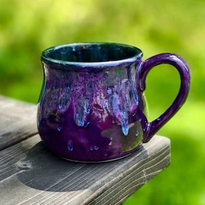 May include: A purple ceramic mug with a blue and green glaze. The mug has a rounded body and a handle. It is sitting on a wooden surface.