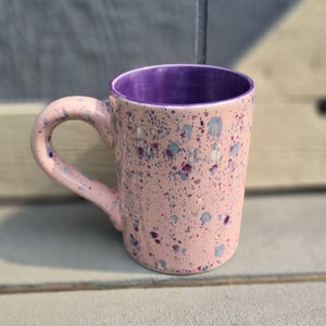 Puede incluir: Una taza de cerámica rosa con interior morado y diseño moteado. La taza tiene un asa redondeada.