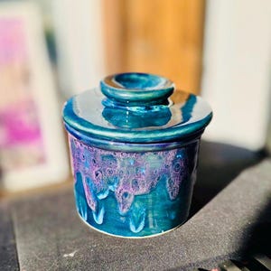 May include: A ceramic butter dish with a lid. The dish is decorated with a blue, purple and white glaze. The glaze has a speckled and drippy effect.