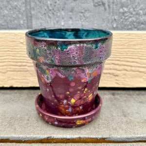 Pode incluir: Um pequeno vaso de cerâmica roxo com um esmalte turquesa e um pires correspondente. O vaso tem um design salpicado, semelhante a uma galáxia, com detalhes dourados e amarelos.
