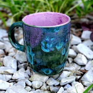 May include: A green and purple ceramic mug with a speckled glaze. The mug has a pink interior and a rounded handle.