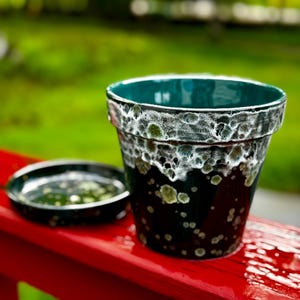 Puede incluir: Una maceta de cerámica y su platillo a juego. La maceta es de color verde azulado oscuro con un borde moteado blanco y verde. El interior es verde azulado. El platillo es verde azulado oscuro con motas verdes.