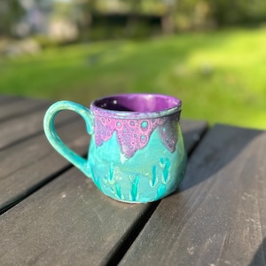 Puede incluir: Una taza de cerámica de color morado y turquesa con un esmalte texturizado y burbujeante y un diseño de escena de montaña.