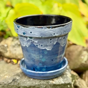 May include: A blue ceramic planter with a matching saucer. The planter has a dark blue interior and a textured, layered design in shades of blue and white. The planter is sitting on a stone surface.