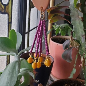 May include: A terracotta-colored clay pendant with a gold chain and purple beads hangs from a gold hook. The pendant is decorated with a gold ring and has a fringe of purple beads. The pendant is hanging in front of a window with stained glass and a potted plant.