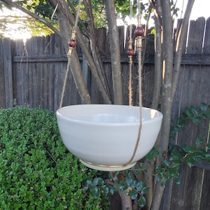 May include: White ceramic hanging planter with a gold rim, suspended from a tree branch by twine with wooden beads.